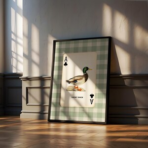 May include: A framed wall art print featuring a playing card design with a green and white gingham background. The card displays an ace of clubs symbol, a mallard duck illustration, and the text LUCKY DUCK printed in black capital letters.