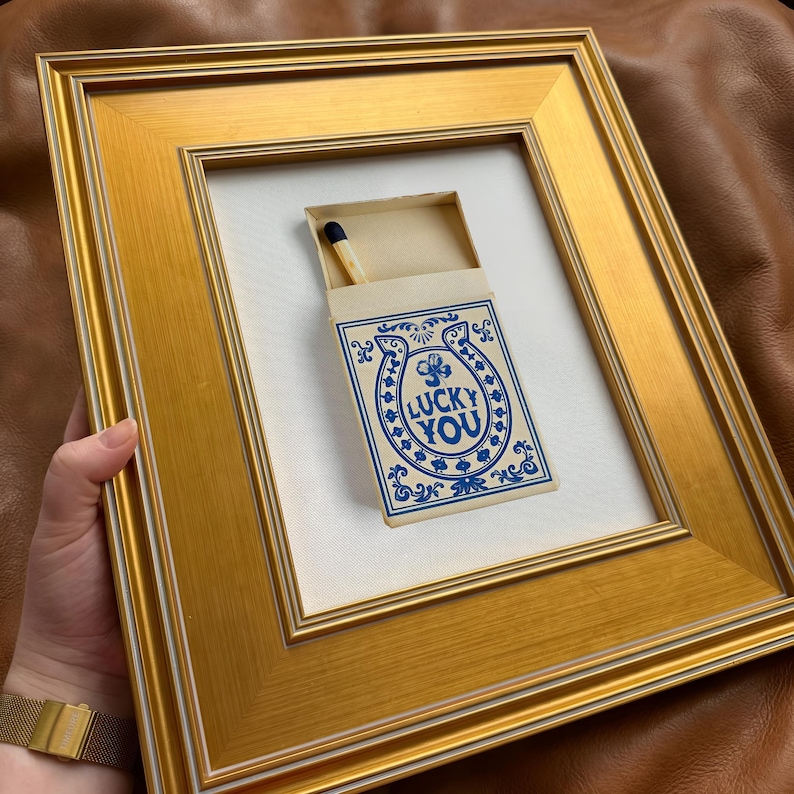 May include: A framed artwork featuring a matchbox with the words "LUCKY YOU" in blue script and a horseshoe design. The matchbox is open, revealing a single match. The frame is gold-colored.
