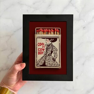 Op de afbeelding: Een ingelijst luciferdoosje kunstwerk. Het zwarte frame omringt een rode achtergrond, met een vintage luciferdoosje. Het luciferdoosje toont een vrouw in een bloemenjurk met de tekst "COOL IT COW BOY". De luciferkoppen zijn rood.