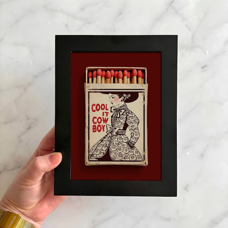 Op de afbeelding: Een ingelijst luciferdoosje kunstwerk. Het zwarte frame omringt een rode achtergrond, met een vintage luciferdoosje. Het luciferdoosje toont een vrouw in een bloemenjurk met de tekst "COOL IT COW BOY". De luciferkoppen zijn rood.