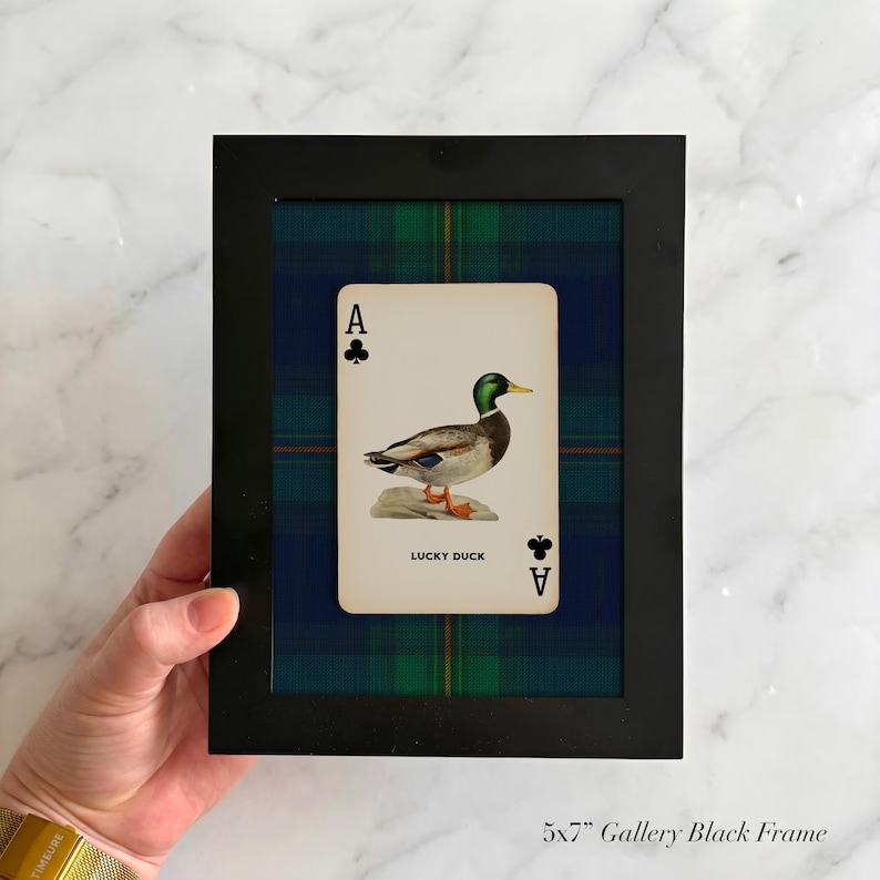 May include: A black framed print featuring a playing card with an illustration of a duck and the text "LUCKY DUCK". The background is a blue, green, and black plaid pattern. The frame is 5x7 inches.