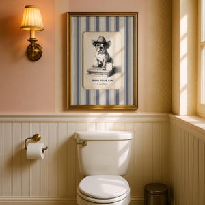 May include: A framed print featuring a bulldog wearing a cowboy hat, with the text "MIND YOUR AIM Cowboy." The artwork is set against a striped background and framed in gold. The print hangs above a white toilet in a bathroom.