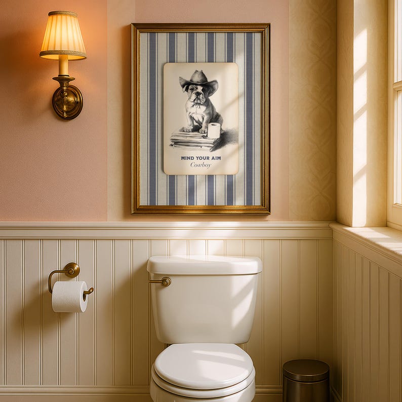 May include: A framed print featuring a bulldog wearing a cowboy hat, with the text "MIND YOUR AIM Cowboy." The artwork is set against a striped background and framed in gold. The print hangs above a white toilet in a bathroom.