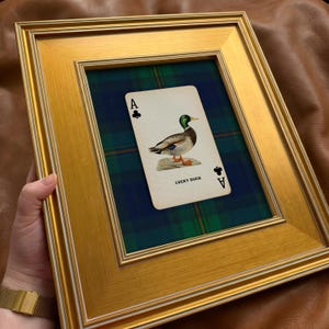 May include: A framed print featuring a playing card with a duck illustration. The card, labeled "LUCKY DUCK," is set against a green and blue plaid background within a gold-colored frame. The ace of clubs is visible.