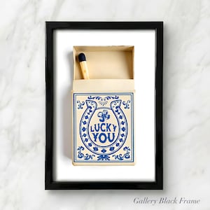 May include: A framed print featuring a matchbox with a blue and cream design. The matchbox has a horseshoe and clover graphic with the words "LUCKY YOU". A single match with a blue tip is visible inside the box.
