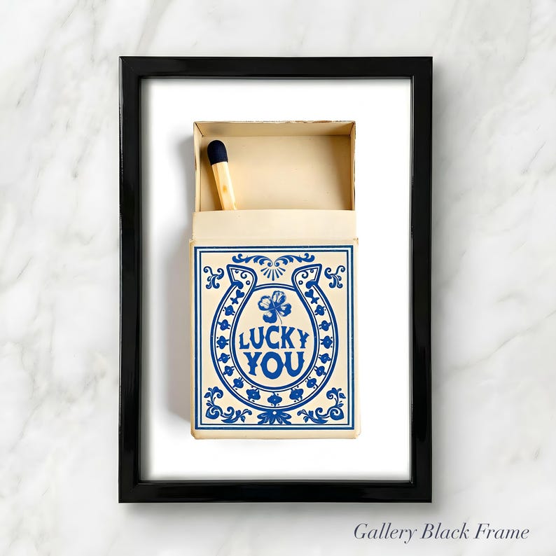 May include: A framed print featuring a matchbox with a blue and cream design. The matchbox has a horseshoe and clover graphic with the words "LUCKY YOU". A single match with a blue tip is visible inside the box.