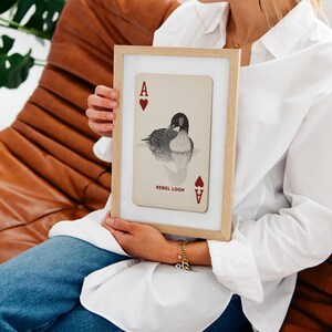 Puede incluir: Una l&aacute;mina enmarcada con un dise&ntilde;o de carta de juego sobre fondo beige, una ilustraci&oacute;n en blanco y negro de un colimbo, corazones rojos en las esquinas y el texto REBEL LOON impreso en rojo. El marco es de madera clara con paspart&uacute; blanco.