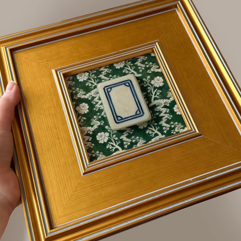 May include: A framed piece featuring a gold-colored wooden frame with a white and silver inner border. Inside, a green floral fabric background showcases a small, rectangular tile with a blue border. The tile is off-white.