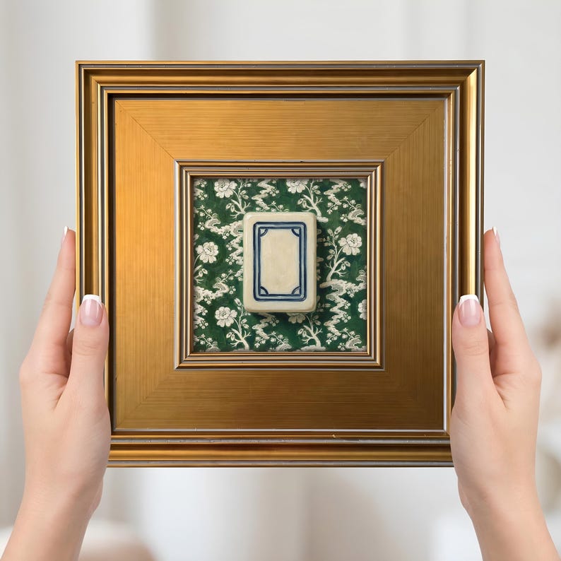 May include: A framed Mahjong tile. The tile is cream-colored with a blue border, set against a green floral background. The frame is gold-colored and square. The image is held by two hands.