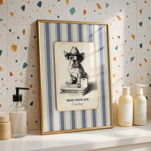 May include: Framed artwork featuring a bulldog wearing a cowboy hat, with the text "MIND YOUR AIM Cowboy." The artwork is set against a blue and white striped background within a gold frame. The image is on a white surface with soap dispensers.