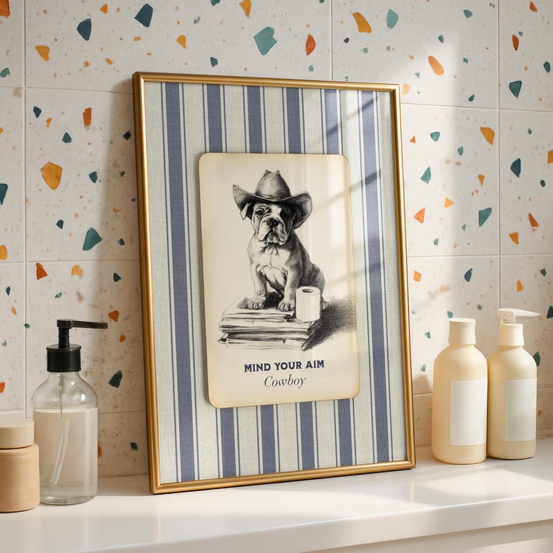 May include: Framed artwork featuring a bulldog wearing a cowboy hat, with the text "MIND YOUR AIM Cowboy." The artwork is set against a blue and white striped background within a gold frame. The image is on a white surface with soap dispensers.