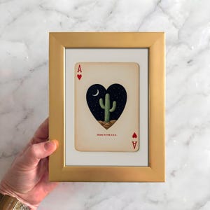 May include: A framed playing card featuring a heart-shaped design with a cactus under a crescent moon and stars. The card is an Ace of Hearts, set in a gold frame. The text "MADE IN THE U.S.A." is visible.