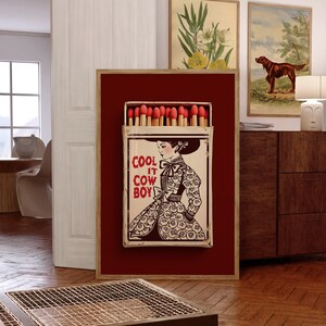 Op de afbeelding: Een ingelijst kunstwerk met een grote luciferdoos met rode koppen. De luciferdoosillustratie toont een vrouw in een patroonjurk met de tekst "COOL IT COW BOY". Het kunstwerk is geplaatst tegen een dieprode achtergrond.