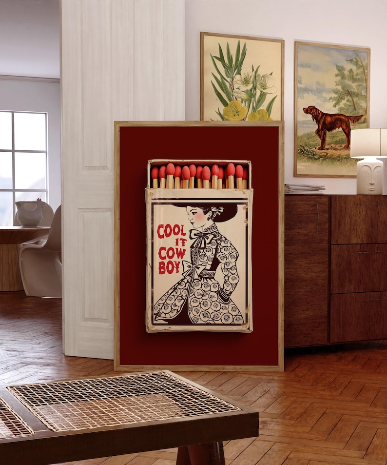 Op de afbeelding: Een ingelijst kunstwerk met een grote luciferdoos met rode koppen. De luciferdoosillustratie toont een vrouw in een patroonjurk met de tekst "COOL IT COW BOY". Het kunstwerk is geplaatst tegen een dieprode achtergrond.