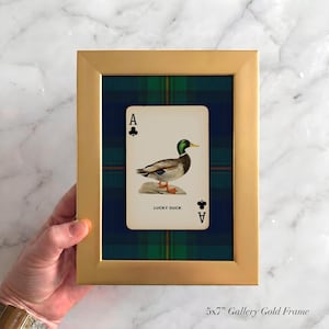 May include: A gold framed print featuring an Ace of Clubs playing card with a duck illustration. The card is set against a blue and green plaid background. The text "LUCKY DUCK" is visible on the card.