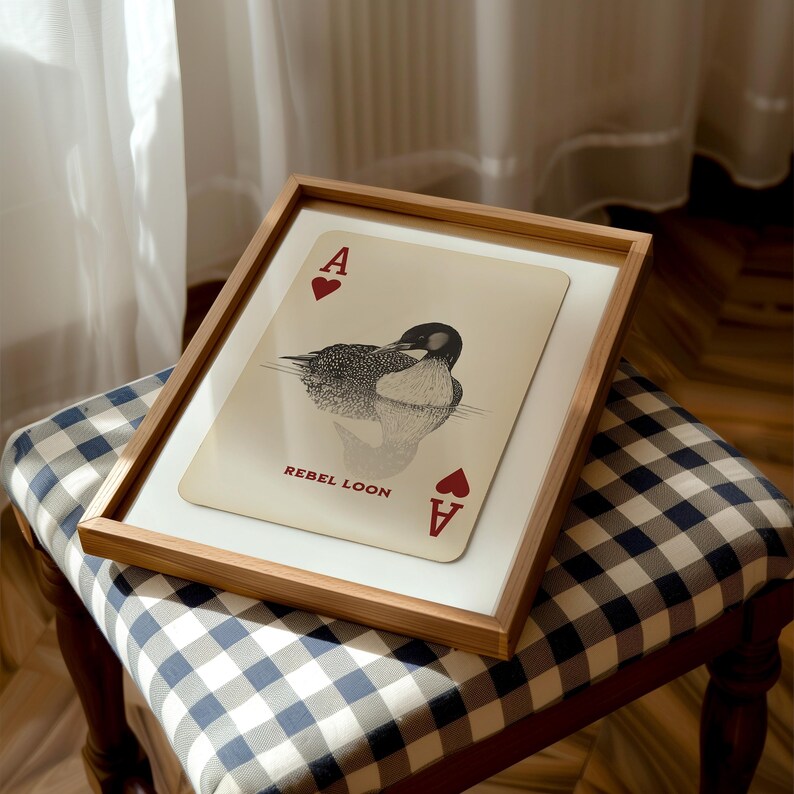 Puede incluir: Una impresi&oacute;n enmarcada con un dise&ntilde;o de carta de as de corazones y una ilustraci&oacute;n en blanco y negro de un colimbo nadando en el agua. El texto REBEL LOON est&aacute; impreso en rojo debajo del ave, con corazones rojos en las esquinas.