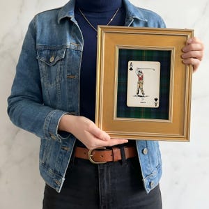 May include: A framed playing card featuring a golfer, labeled "ACE IT." The card is set against a green and blue plaid background within a gold-colored frame. The frame is rectangular and has a simple, elegant design.