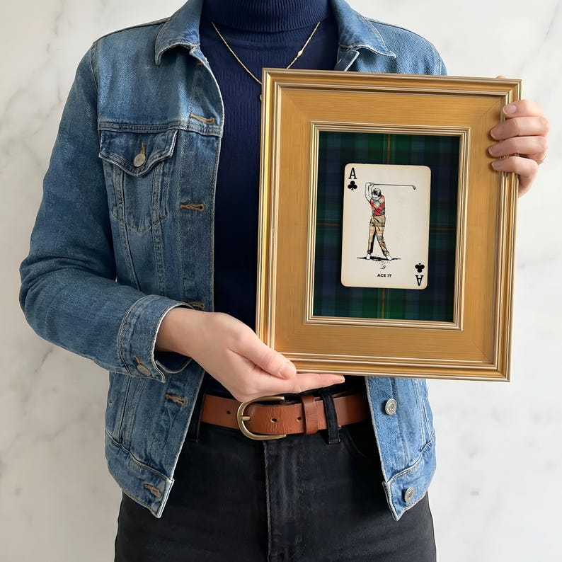 May include: A framed playing card featuring a golfer, labeled "ACE IT." The card is set against a green and blue plaid background within a gold-colored frame. The frame is rectangular and has a simple, elegant design.