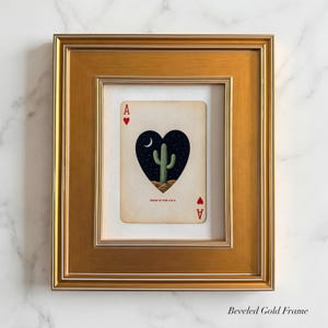 May include: Framed playing card art featuring the Ace of Hearts. The card displays a heart-shaped night scene with a cactus, moon, and stars. The card is framed in a gold beveled frame.