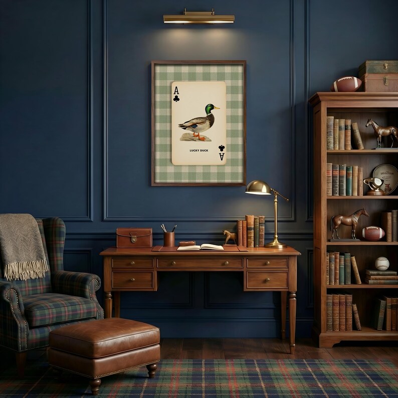 May include: A framed wall art print featuring an Ace of Clubs playing card with a mallard duck illustration and the text Lucky Duck, centered above a wooden desk with drawers, a brass lamp, and a bookshelf filled with books and figurines.