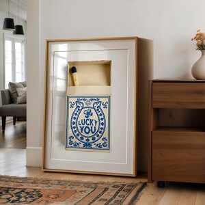 May include: Framed artwork featuring a large matchbox design with the text "LUCKY YOU" in blue. The matchbox is open, revealing a single match. The artwork is in a wooden frame and leans against a white wall.