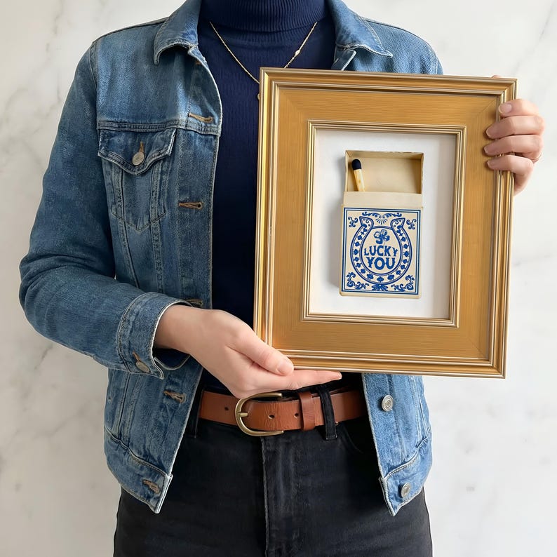 May include: A framed artwork featuring a matchbox with a blue and white design and the words "LUCKY YOU." The matchbox is open, revealing a single match. The frame is gold-colored.