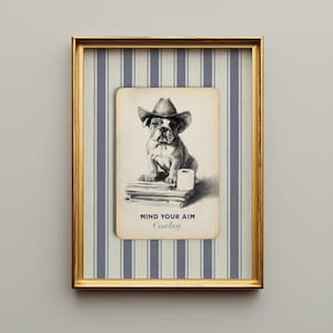 An illustration of an English Bulldog wearing a cowboy hat and sitting on a stack of newspapers next to a roll of toilet paper. Below it says "Mind you aim, Cowboy" in rustic lettering. The design is set on a vintage field notecard and lays on a navy blue and cream striped washed linen background. The whole design comes printed on a single layer. Shown here framed in a vintage beveled gold frame.