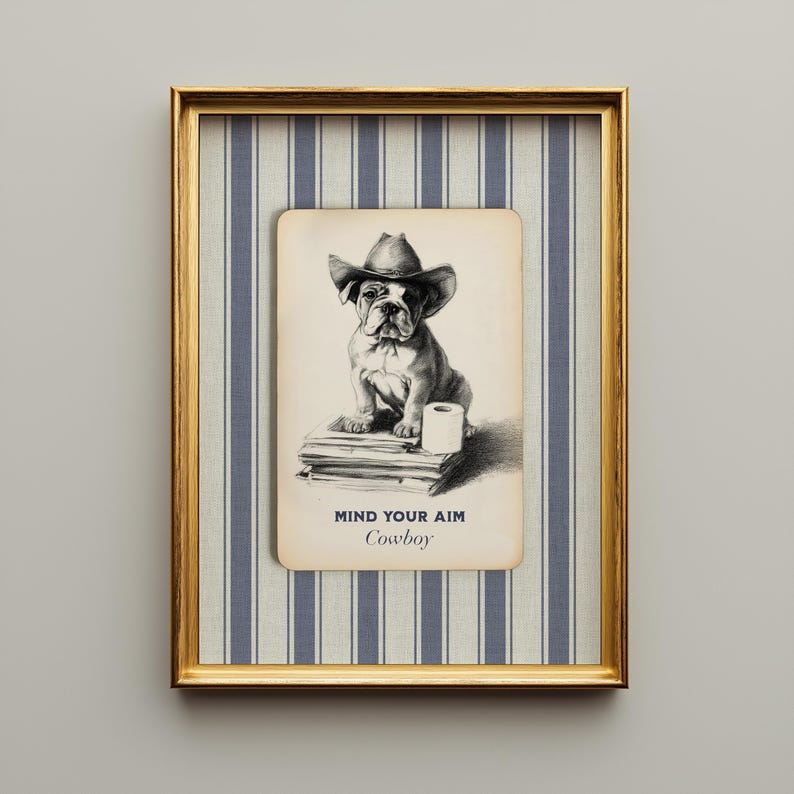 An illustration of an English Bulldog wearing a cowboy hat and sitting on a stack of newspapers next to a roll of toilet paper. Below it says "Mind you aim, Cowboy" in rustic lettering. The design is set on a vintage field notecard and lays on a navy blue and cream striped washed linen background. The whole design comes printed on a single layer. Shown here framed in a vintage beveled gold frame.