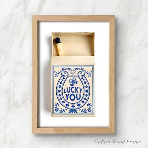 May include: A framed print featuring a matchbox with a blue and white design. The matchbox has the words "LUCKY YOU" inside a horseshoe and clover. A single match with a black tip is visible.
