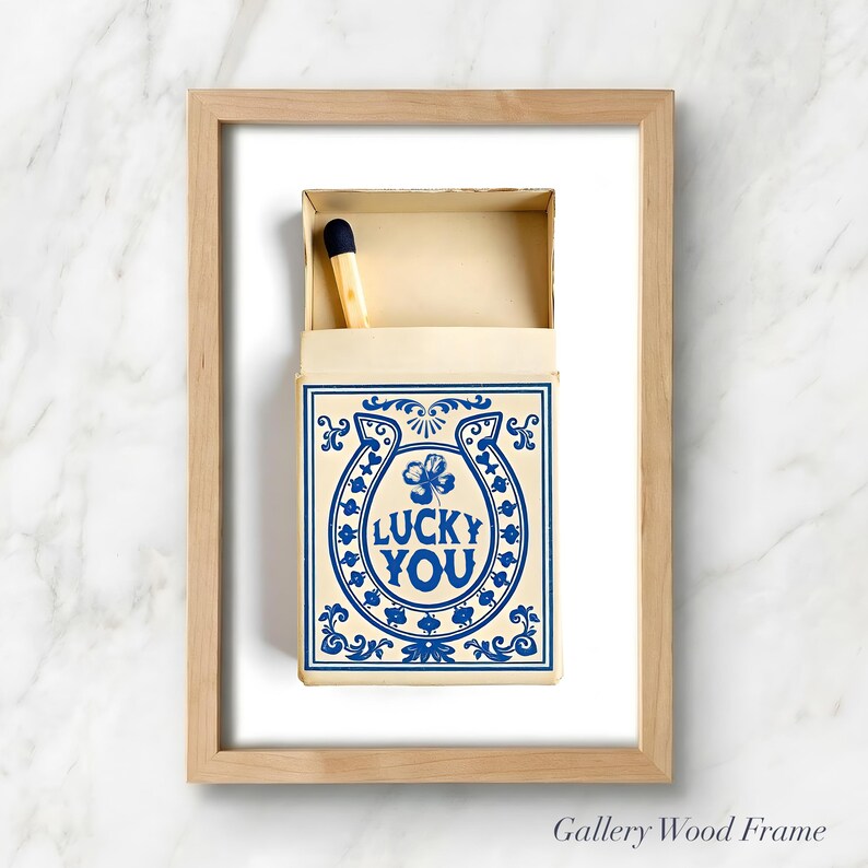 May include: A framed print featuring a matchbox with a blue and white design. The matchbox has the words "LUCKY YOU" inside a horseshoe and clover. A single match with a black tip is visible.