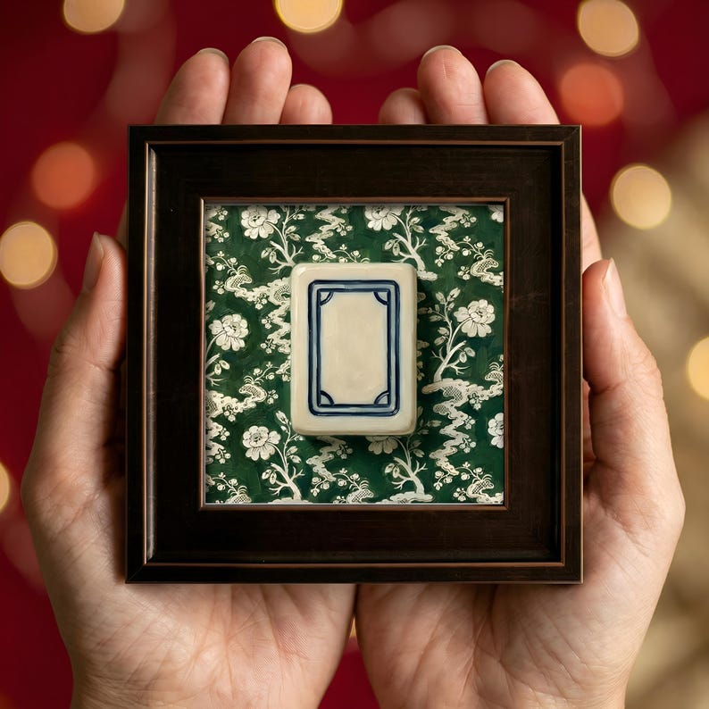 A framed Mahjong tile with a cream-colored center and blue border, set against a green floral chinoiserie patterned background. The dark brown frame is held in open hands, with a blurred red bokeh background. Showing mini painting size and ideal mother's day gift or game night hostess gift keepsake.