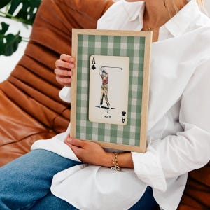 K&ouml;nnte beinhalten: Ein gerahmter Druck mit einem Spielkartendesign auf gr&uuml;n-wei&szlig; kariertem Hintergrund. Die Karte zeigt die Illustration eines Golfers in rotem Hemd und karierter Hose, mit dem Text ACE IT und dem Kreuz-Symbol in den Ecken.