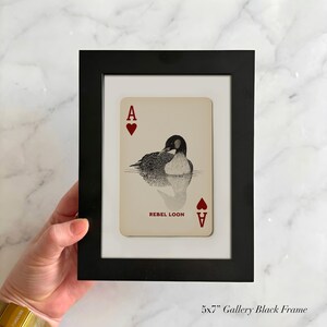 Puede incluir: Un marco de galer&iacute;a negro de 13x18 cm que contiene una carta de juego con fondo crema. La carta presenta un s&iacute;mbolo de as de corazones rojo en las esquinas, una ilustraci&oacute;n en blanco y negro de un colimbo en el centro y el texto REBEL LOON.