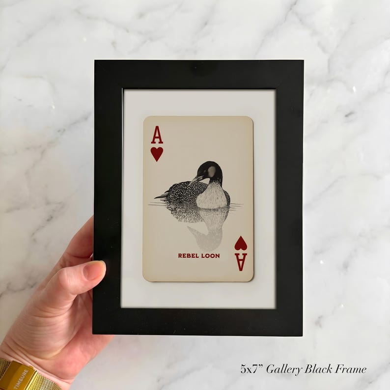 Puede incluir: Un marco de galer&iacute;a negro de 13x18 cm que contiene una carta de juego con fondo crema. La carta presenta un s&iacute;mbolo de as de corazones rojo en las esquinas, una ilustraci&oacute;n en blanco y negro de un colimbo en el centro y el texto REBEL LOON.