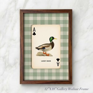 May include: A framed wall art piece featuring a playing card design with a mallard duck illustration, the text LUCKY DUCK, and club symbols. The card is set against a green and white gingham background within a 12-inch by 18-inch walnut wood frame.