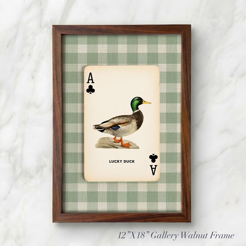 May include: A framed wall art piece featuring a playing card design with a mallard duck illustration, the text LUCKY DUCK, and club symbols. The card is set against a green and white gingham background within a 12-inch by 18-inch walnut wood frame.