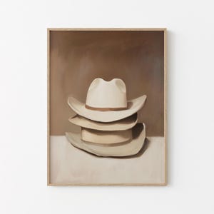 Puede incluir: Una impresión artística enmarcada que presenta una pila de tres sombreros de vaquero. El sombrero superior es blanco con una banda marrón, y los otros dos sombreros son de color beige. El fondo es un degradado de marrón y beige.