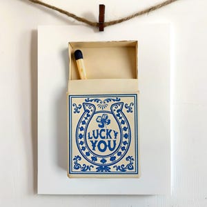 May include: A matchbox with a blue horseshoe and clover design, with the words "LUCKY YOU" in blue. The matchbox is open, revealing a single match with a blue tip. The matchbox is mounted on a white background and suspended by a clothes pin and twine.