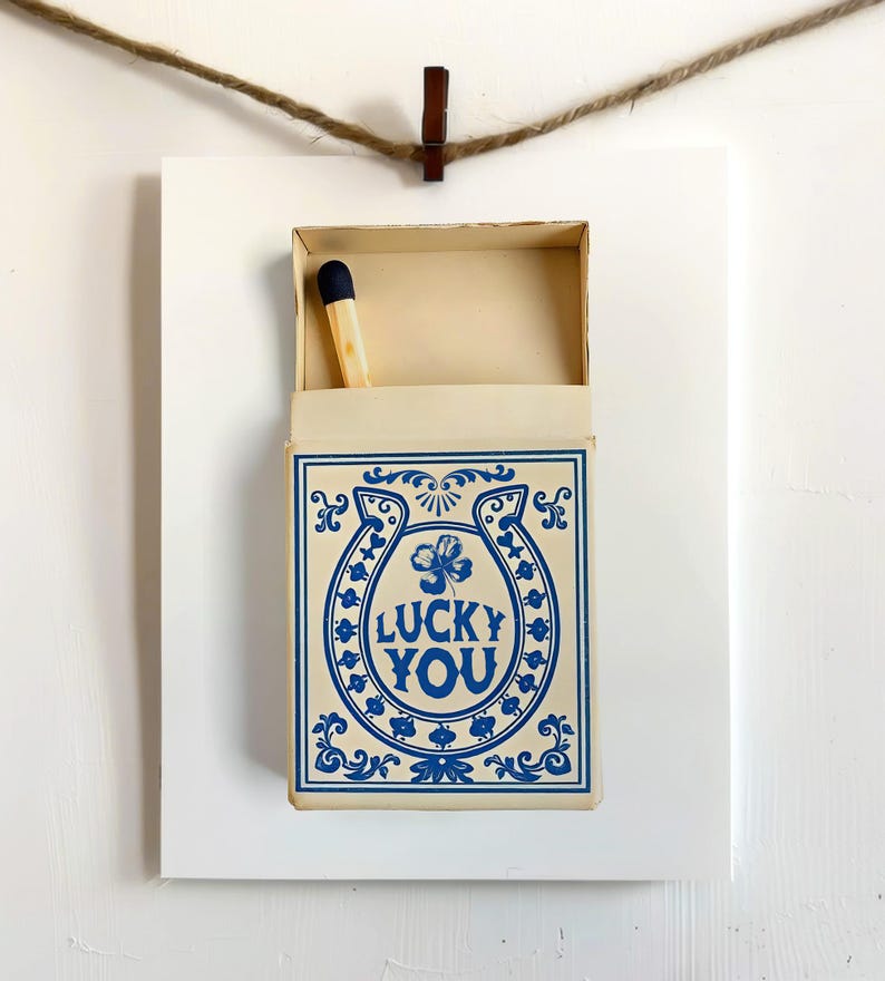 May include: A matchbox with a blue horseshoe and clover design, with the words "LUCKY YOU" in blue. The matchbox is open, revealing a single match with a blue tip. The matchbox is mounted on a white background and suspended by a clothes pin and twine.