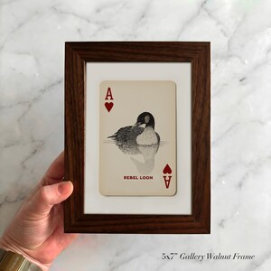 Puede incluir: Un marco de galer&iacute;a de nogal de 13x18 cm que contiene una carta de juego con una ilustraci&oacute;n de un colimbo sobre el agua. La carta muestra la letra A y un s&iacute;mbolo de coraz&oacute;n rojo en las esquinas, con el texto REBEL LOON impreso debajo.