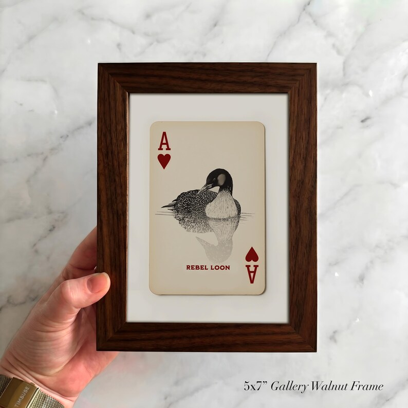 Puede incluir: Un marco de galer&iacute;a de nogal de 13x18 cm que contiene una carta de juego con una ilustraci&oacute;n de un colimbo sobre el agua. La carta muestra la letra A y un s&iacute;mbolo de coraz&oacute;n rojo en las esquinas, con el texto REBEL LOON impreso debajo.