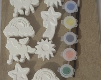 May include: A craft kit featuring white plaster shapes of unicorns, stars, rainbows, and suns. Includes six paint pots in various colors and a small paintbrush. The kit is designed for painting and creative activities.