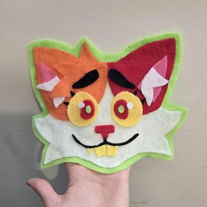 Pode incluir: Uma face de gato de feltro feita à mão em laranja, vermelho, branco, amarelo e verde. O gato tem olhos amarelos grandes com pupilas vermelhas, sobrancelhas pretas e uma boca sorridente. A face do gato é delineada em verde.