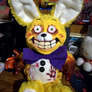 May include: A yellow plush rabbit with large ears, a purple bow tie, and a wide, toothy grin. The rabbit has blood-like stains on its chest and arms, and its eyes are green with red outlines. The toy has black buttons and is a unique, handmade item.