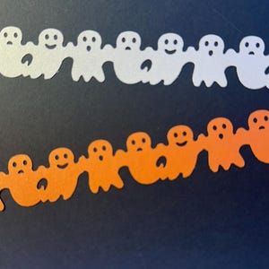 Puede incluir: Dos filas de recortes de fantasmas de papel sobre un fondo oscuro. La fila superior presenta fantasmas blancos con diversas expresiones faciales. La fila inferior tiene fantasmas naranjas con expresiones similares. Decoraciones de Halloween.