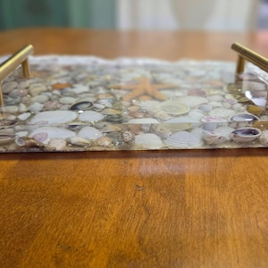 May include: A rectangular serving tray with a clear resin top filled with seashells, sand, and a starfish. The tray has gold-colored handles on each end. The tray is on a wooden surface.