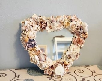 May include: A heart-shaped mirror with a frame made of seashells. The mirror is surrounded by a variety of seashells, including sand dollars, starfish, and other shells.
