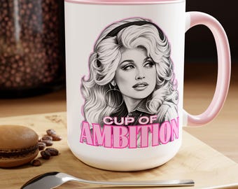 Puede incluir: Una taza de cerámica blanca con interior y asa de color rosa claro. El frente presenta una ilustración en blanco y negro de una mujer con cabello rubio voluminoso y pendientes de aro, con el texto rosa "CUP OF AMBITION" impreso debajo.