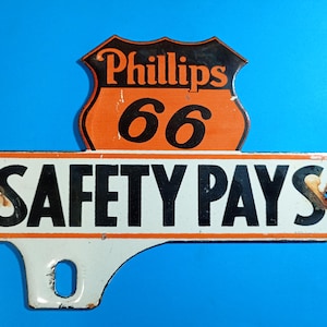 May include: A vintage metal sign featuring the Phillips 66 logo in orange and black at the top, with a white rectangular section below displaying the black text SAFETY PAYS. The sign has visible rust spots near the mounting holes.