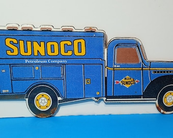 Puede incluir: Un letrero de metal azul con forma de camión de servicio con detalles en amarillo. El texto SUNOCO aparece en letras amarillas grandes en el lateral, con Petroleum Company debajo. Un pequeño logotipo en forma de diamante dice BLUE SUNOCO con flechas rojas.
